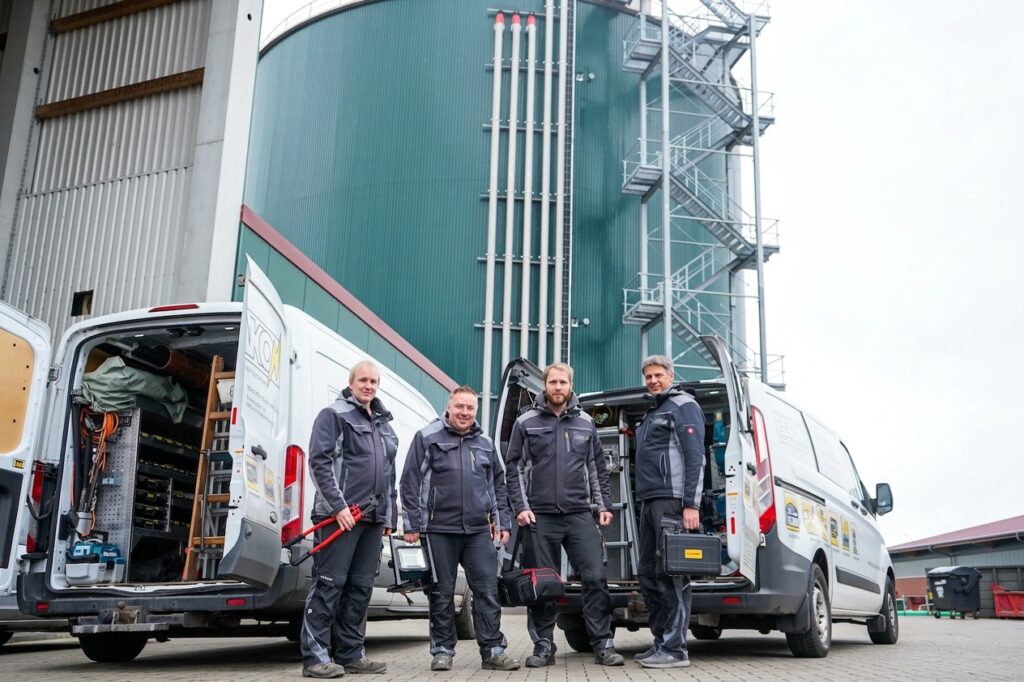 Vier Männer mit neutralem Ausdruck stehen in dunkler Arbeitskleidung vor Bauwerk